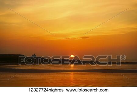 Stock Photograph - The golden sun sinking on the horizon with a silhouette of the coastline and an orange sky reflected in the water; Bamburgh, Northumberland, England. Fotosearch