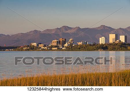Stock Photograph - The setting sun reflects off of office buildings in downtown Anchorage, seen from Earthquake Park, golden grass along the shoreline in the foreground, the Chugach Mountains in the background, South-central Alaska; Anchorage, Alaska, United States of.... Fotosearch