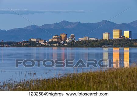 Stock Photograph - The setting sun reflects off of office buildings in downtown Anchorage, seen from Earthquake Park, golden grass along the shoreline in the foreground, the Chugach Mountains in the background, South-central Alaska; Anchorage, Alaska, United States of.... Fotosearch