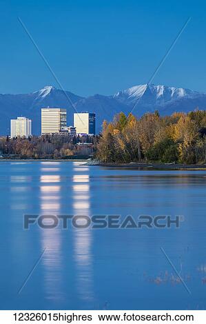 Stock Photograph - The setting sun reflects off of office buildings in downtown Anchorage, seen from Earthquake Park, autumn coloured trees along the shoreline, the Chugach Mountains in the background, South-central Alaska; Anchorage, Alaska, United States of America. Fotosearch