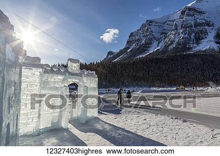 Stock Photograph - The sun shines behind an ice castle constructed on frozen Lake Louise in Banff National Park with figure skaters skating on the lake in the background; Lake Louise, Alberta, Canada. Fotosearch
