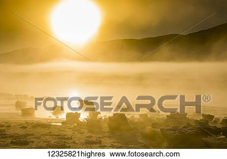 Stock Photograph - The sun shines through low altitude fog cast in warm light along Turnagain Arm and the Seward Highway in winter, sea ice covering the ocean in the foreground, the Chugach Moutains revealed in the background, South-central Alaska; Alaska, United States.... Fotosearch