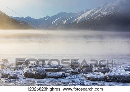 Stock Photograph - The sun shines through low altitude fog cast in warm light along Turnagain Arm and the Seward Highway, sea ice covering the ocean in the foreground, the Kenai Moutains revealed in the background, South-central Alaska; Alaska, United States of America. Fotosearch
