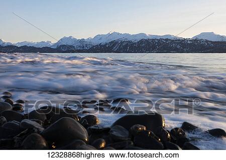 Stock Photograph - The surf rolling into rocks on the shore with snow covered mountain range in the background; Alaska, United States of America. Fotosearch