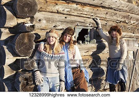 Stock Photograph - Three girls standing against a log wall wearing fur hats; Homer, Alaska, United States of America. Fotosearch