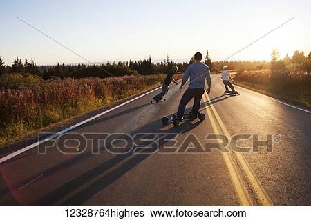 Stock Photograph - Three young men skateboarding down a road at sunset; Homer, Alaska, United States of America. Fotosearch