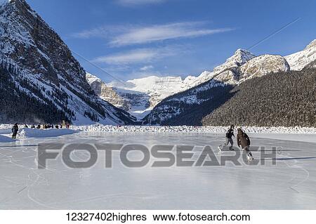 Stock Photograph - Tourists enjoy ice skating on the frozen lake on a sunny day with this scenic mountain view; Lake Louise, Alberta, Canada. Fotosearch