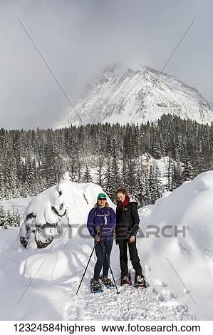 Stock Photograph - Two female snowshoers on a snow covered trail with snow covered mountain in the background; Kananaskis Country, Alberta, Canada. Fotosearch