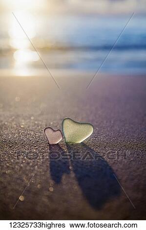 Stock Photograph - Two glass hearts in the sand on the beach at the water's edge; Honolulu, Oahu, Hawaii, United States of America. Fotosearch