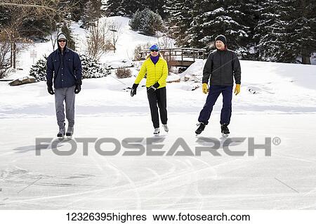 Stock Photograph - Two males and one female skating on pond with snow covered bridge in the background; Calgary, Alberta, Canada. Fotosearch