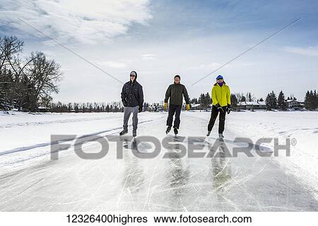 Stock Photograph - Two males and one female skating on freshly groomed ice on pond with houses in the background; Calgary, Alberta, Canada. Fotosearch