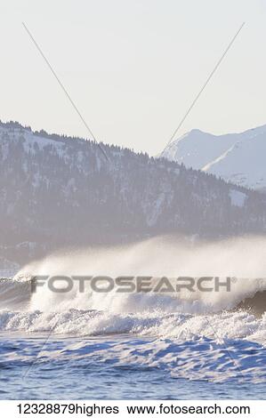 Stock Photograph - Waves splashing and forming a mist over the ocean with snow covered forest and mountains in the background; Alaska, United States of America. Fotosearch