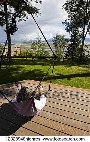 Stock Photograph - Woman relaxing in hammock on wooden deck with view of the ocean and mountains; Alaska, United States of America. Fotosearch