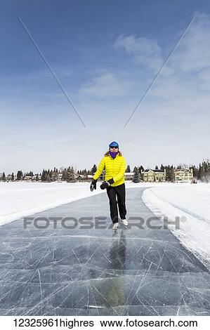 Stock Photograph - Woman skating on freshly groomed ice on lake with houses in the background and blue sky; Calgary, Alberta, Canada. Fotosearch