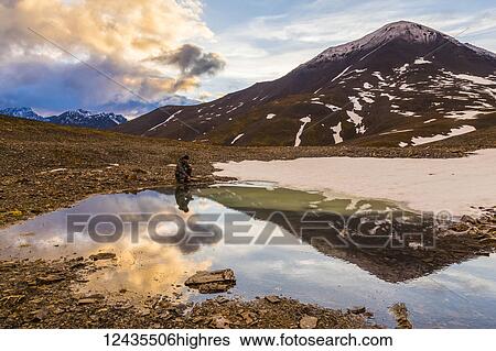 Stock Photograph - A backpacker pauses for rest beside a puddle while crossing over an unnamed pass in the Brooks Range under the midnight sun at 12: 14 a. m.; Alaska, United States of America. Fotosearch