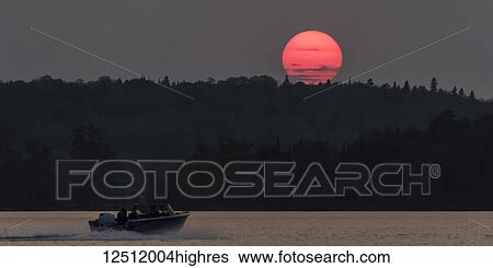 A bright pink sun sinking below a silhouetted hill and forest and a motorboat splashes on a lake; Kenora, Ontario, Canada View Large Photo Image Stock Photograph - A bright pink sun sinking below a silhouetted hill and forest and a motorboat splashes on a lake; Kenora, Ontario, Canada. Fotosearch