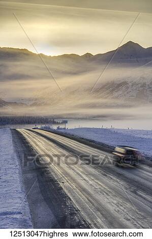 Stock Photograph - A car drives down a road in a snow-covered landscape, the sun rising above a mountainous ridge-line in the background, fog obscuring the skyline in winter, Turnagain Pass, Kenai Peninsula, South-central Alaska; Alaska, United States of America. Fotosearch