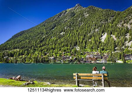 Stock Photograph - A couple enjoys the view on a bench at Champex Lake shore under blue sky, mountain and resorts are in the background; Champex, Valais, Switzerland. Fotosearch