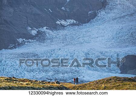 Stock Photograph - A Couple Standing In Front Of A Glacier Along The South Coast Of Iceland, Admiring Nature At Its Best; Iceland. Fotosearch