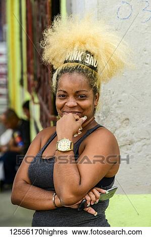 Stock Photograph - A Cuban woman with a unique blond hairstyle stands holding her smart phone and posing for the camera; Havana, Cuba. Fotosearch