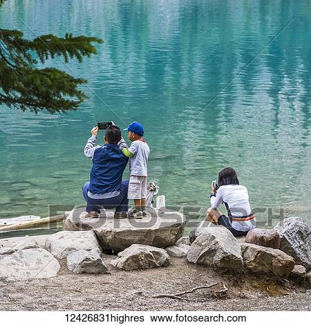 Stock Photograph - A family standing on rocks on the shore of Moraine Lake taking pictures of the tranquil, turquoise water; Field, Alberta, Canada. Fotosearch