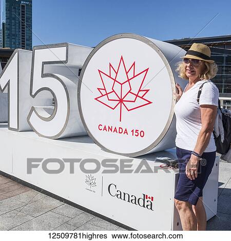 Stock Photograph - A female tourist poses beside a sculpture commemorating Canada's 150th anniversary of confederation; Vancouver, British Columbia, Canada. Fotosearch
