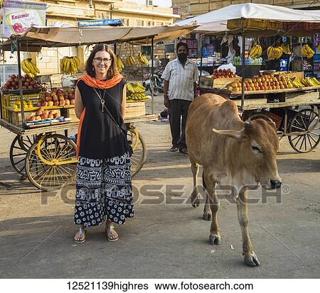 Stock Photograph - A female tourist stands on a street with a cow and food vendors; Jaisalmer, Rajasthan, India. Fotosearch