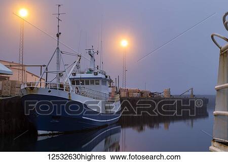 Stock Photograph - A fishing vessel sits idle in the late night fog at the docks of Djupivogur village, Eastern Iceland in summer solstice; Djupivogur, Iceland. Fotosearch