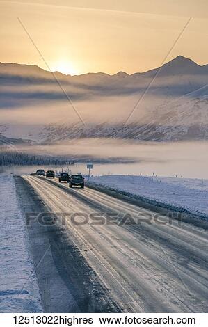 Stock Photograph - A foggy sunset illuminates cars driving down a snow-covered Seward Highway, the Kenai Mountains in the background, Kenai Peninsula, South-central Alaska in winter; Alaska, United States of America. Fotosearch