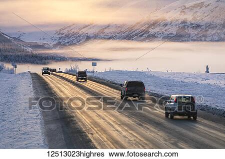 Stock Photograph - A foggy sunset illuminates cars driving down a snow-covered Seward Highway, the Kenai Mountains in the background, Kenai Peninsula, South-central Alaska in winter; Alaska, United States of America. Fotosearch