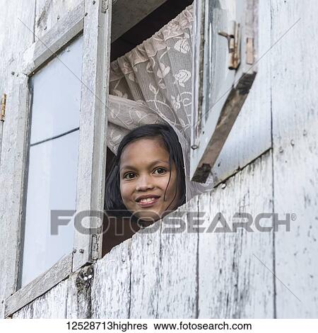 Stock Photograph - A girl looks out an open window and smiles as she looks down at the camera; Sikkim, India. Fotosearch