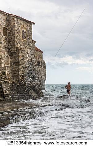 Stock Photograph - A man in a swimsuit stands in the water at the shoreline looking out at the Adriatic Sea; Budva, Opstina Budva, Monte. Fotosearch