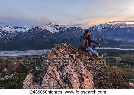 Stock Photograph - A man observes a tranquil sunset from an alpine perch in the Alaska Range high above the Delta River; Alaska, United States of America. Fotosearch