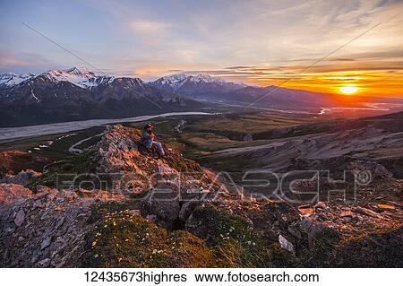 Stock Photograph - A man observes a tranquil sunset from an alpine perch high above the Delta River in the Alaska Range; Alaska, United States of America. Fotosearch
