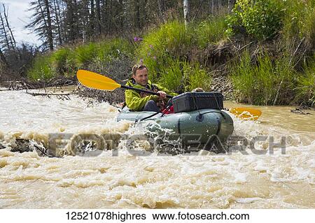 Stock Photograph - A man pack rafting on Jarvis Creek floats over a submerged log, Interior Alaska; Alaska, United States of America. Fotosearch