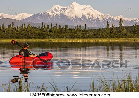 Stock Photograph - A man paddles a pack raft across Donnelly Lake with Mt. Hayes towering in the distance; Alaska, United States of America. Fotosearch
