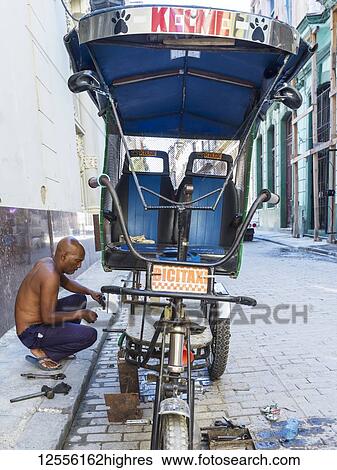 Stock Photograph - A man repairs his cycle rickshaw on the side of the street; Havana, Cuba. Fotosearch