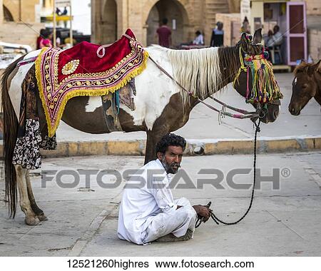 Stock Photograph - A man sits on the ground with his decorated horse standing beside him; Jaisalmer, Rajasthan, India. Fotosearch