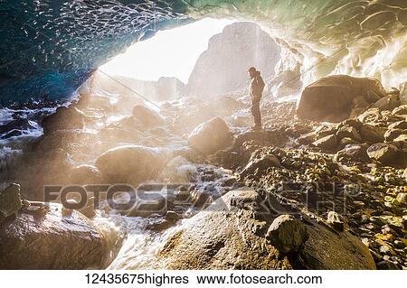 Stock Photograph - A man stands at the entrance of an ice cave bathed in warm sunlight and mist, Wrangell-St. Elias National Park; Alaska, United States of America. Fotosearch