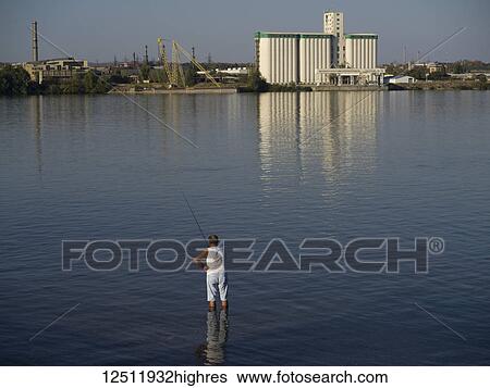 Stock Photograph - A man stands in knee-deep water in the Danube river fishing in his shorts and sleeveless shirt; Kladovo, Bor District, Serbia. Fotosearch
