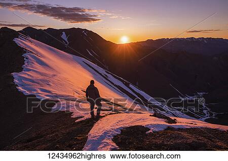 Stock Photograph - A man stands silhouetted on a ridge as the sun sets behind Polychrome Mountain in Denali National Park in early summer; Alaska, United States of America. Fotosearch