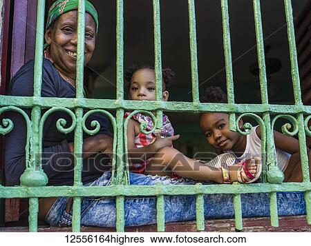 Stock Photograph - A mother with two young daughters sits looking out bars from their home; Havana, Cuba. Fotosearch