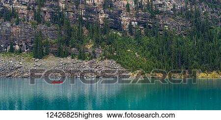 Stock Photograph - A red canoe in Moraine Lake with a cliff of the Canadian Rocky Mountains along the shoreline; Lake Louise, Alberta, Canada. Fotosearch