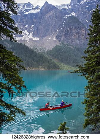 Stock Photograph - A red canoe in Moraine Lake with a cliff of the Canadian Rocky Mountains along the shoreline; Lake Louise, Alberta, Canada. Fotosearch