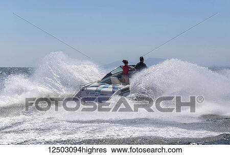 Stock Photograph - A speedboat hits a large ocean wave sending out a huge splash as the driver and passenger stand at the bow looking ahead; Vancouver, British Columbia, Canada. Fotosearch