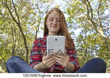 Stock Photograph - A teenage girl wearing jeans and a plaid shirt sits on a boulder using her smart phone, Woodbine Beach; Toronto, Ontario, Canada. Fotosearch