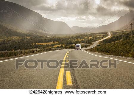 Stock Photograph - A vehicle with trailer heads down a road leading through fields and mountains under a cloudy sky; Newfoundland, Canada. Fotosearch