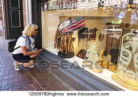 Stock Photograph - A woman crouches to look at a totem sculpture in a shop's window display; Vancouver, British Columbia, Canada. Fotosearch