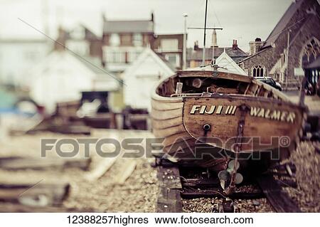 Stock Photograph - A wooden motorboat sits on the shingle beach with buildings in the background; England. Fotosearch