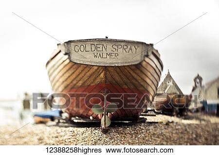 Stock Photograph - A wooden motorboat sits on the shingle beach with buildings in the background; England. Fotosearch